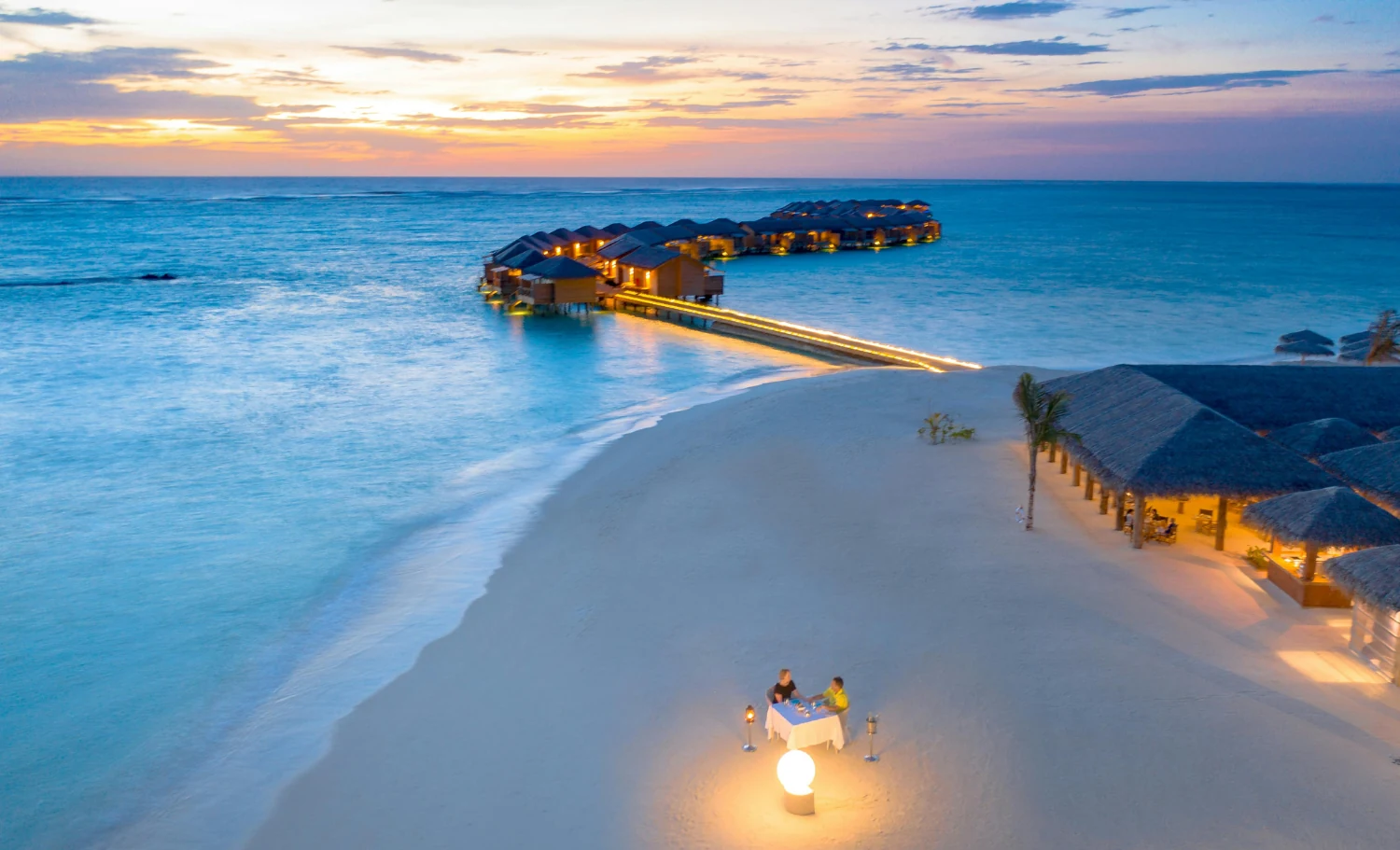 Cena privada en la playa frente a villas sobre el agua durante una luna de miel